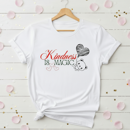 White t-shirt with 'Kindness is Magic' text and bear graphic on a wooden surface with decorative elements.